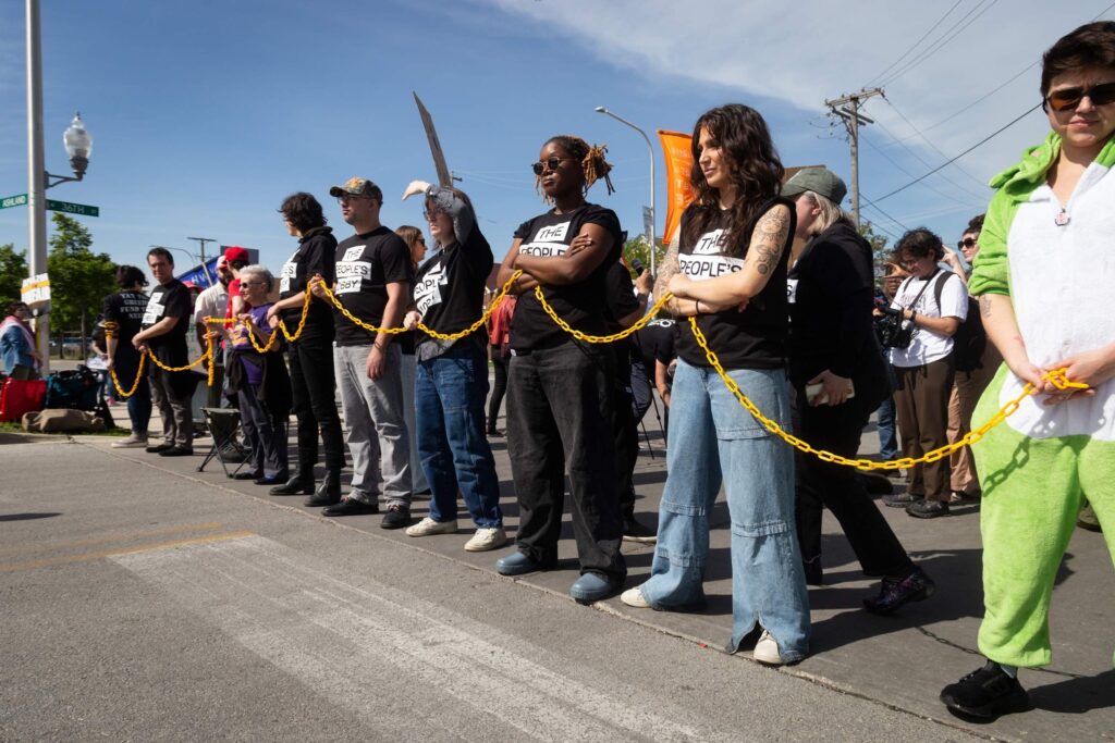 People standing in line, side by side, facing Amazone warehouse linked by yellow chain.