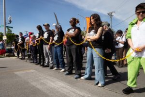 People standing in line, side by side, facing Amazone warehouse linked by yellow chain.