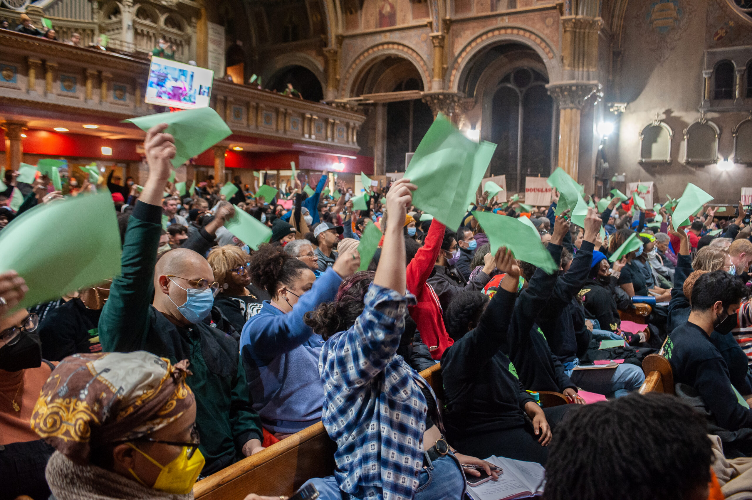 A large group of people in an ornate hall raise green papers in the air during a meeting or assembly.
