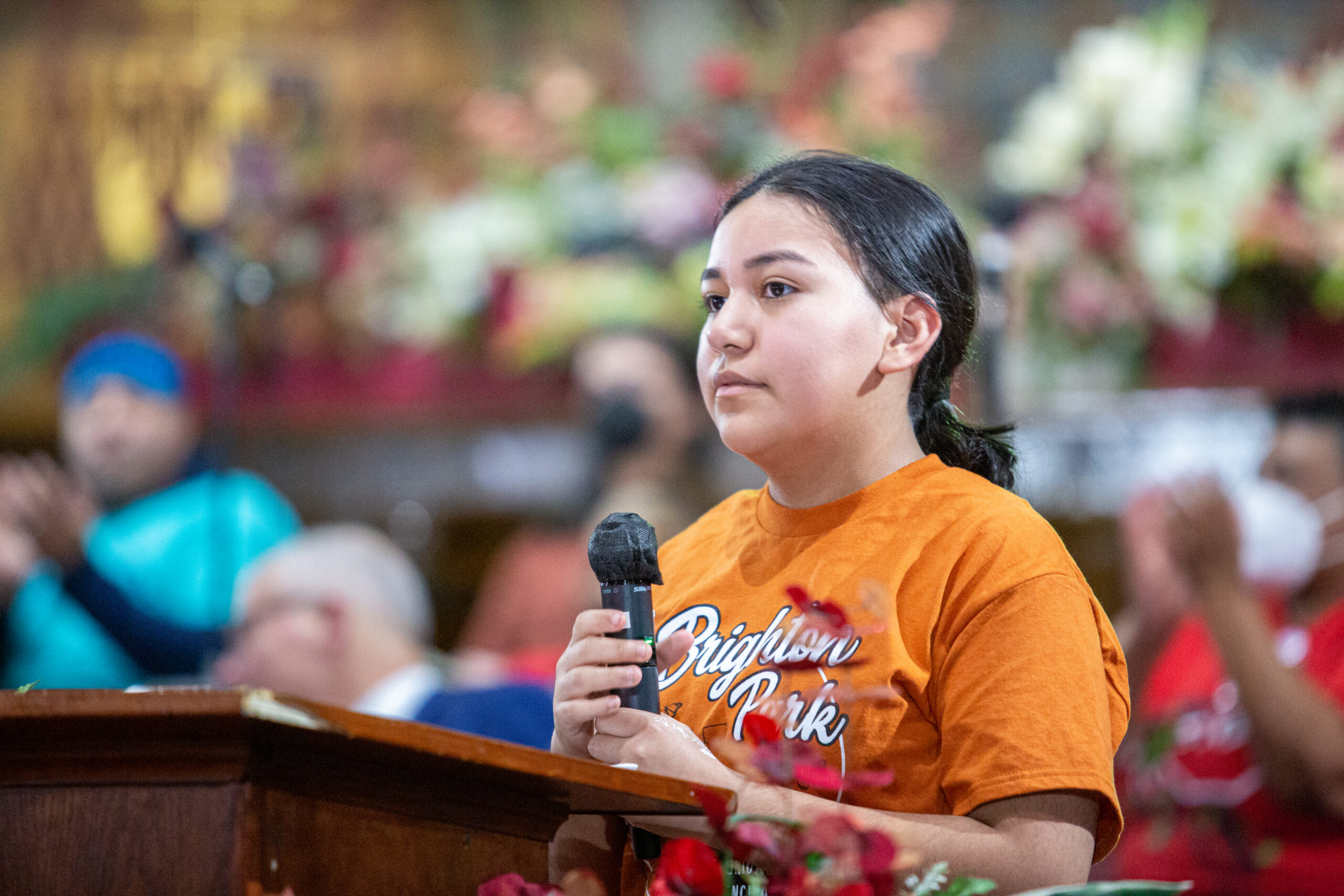 A young person in an orange shirt stands at a podium holding a microphone, with people and floral decorations visible in the background.