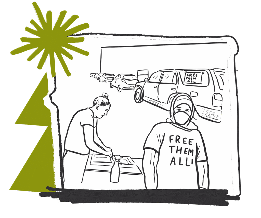 A person wearing a "Free Them All" shirt and face mask stands in front of another person working at a table, with cars and a "Free Them All" sign visible in the background.