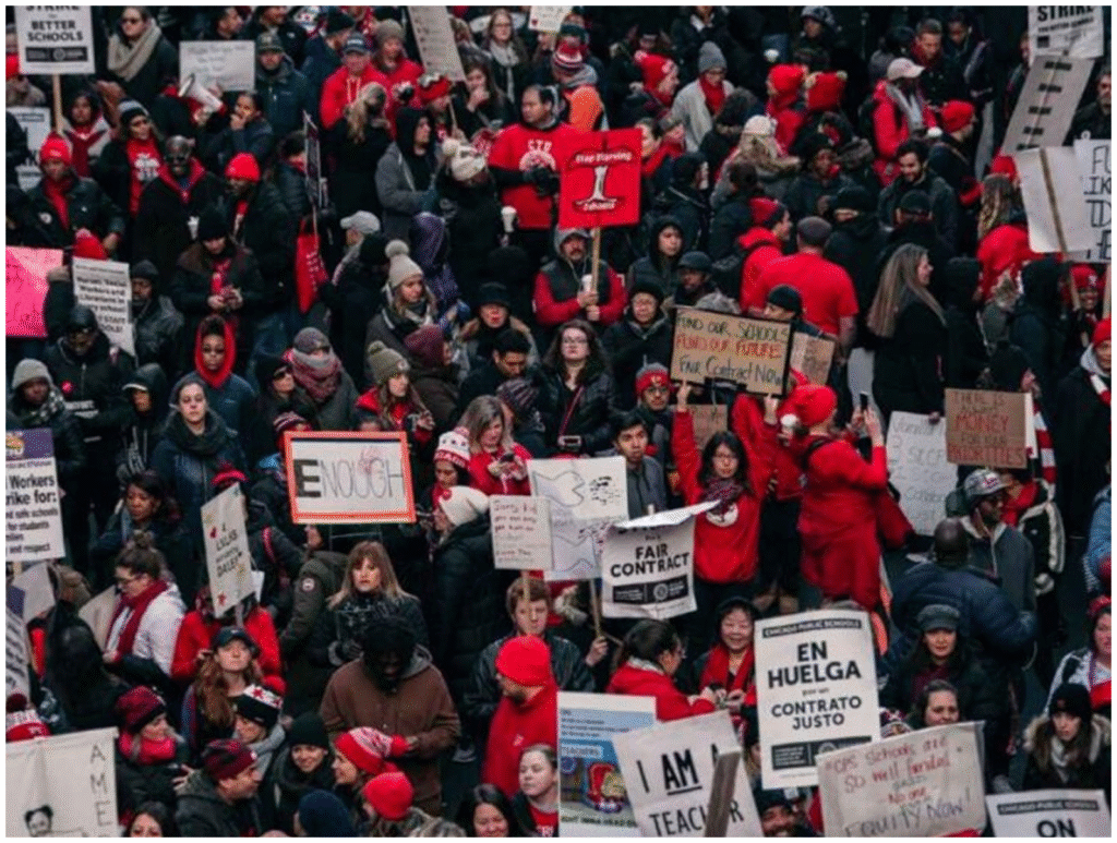 A large crowd of people, many wearing red, hold signs with messages about fair contracts, strikes, and education during a protest or demonstration.