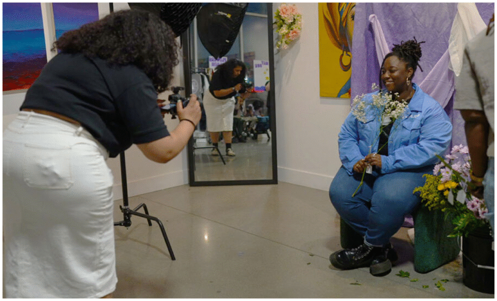 A photographer takes a picture of a seated person holding flowers in a studio with art, flowers, and a mirror reflecting the scene.