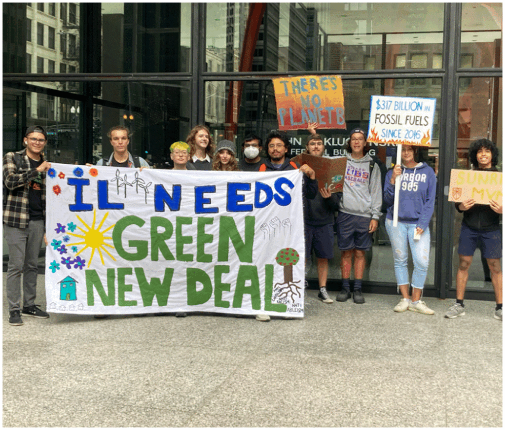 A group of people stand outside holding signs advocating for climate action, including a large banner reading “IL Needs Green New Deal.”.