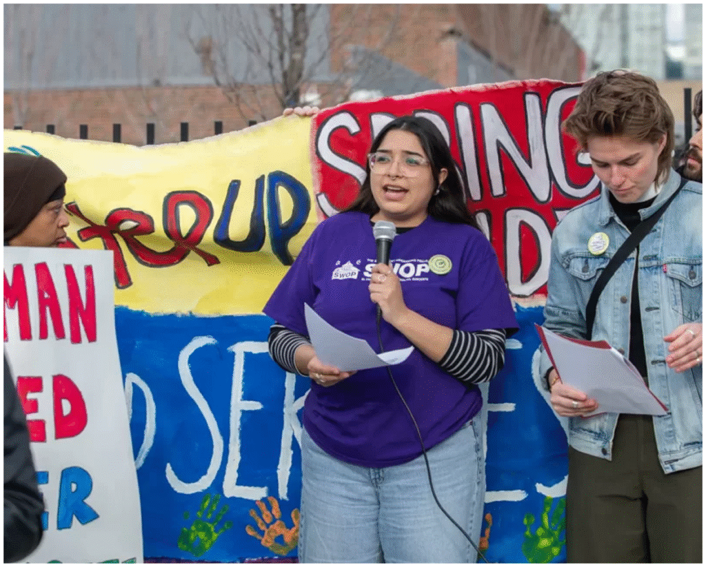 Three people stand in front of a colorful protest sign. One woman speaks into a microphone while holding papers; the other two read from sheets.