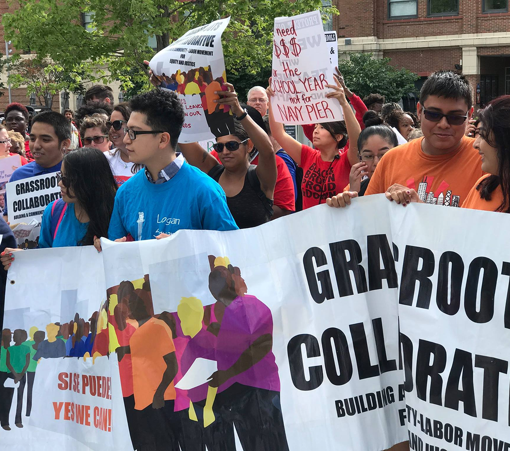 A group of people participate in a protest, holding banners and signs advocating for grassroots collaboration and increased pay for Navy Pier workers.