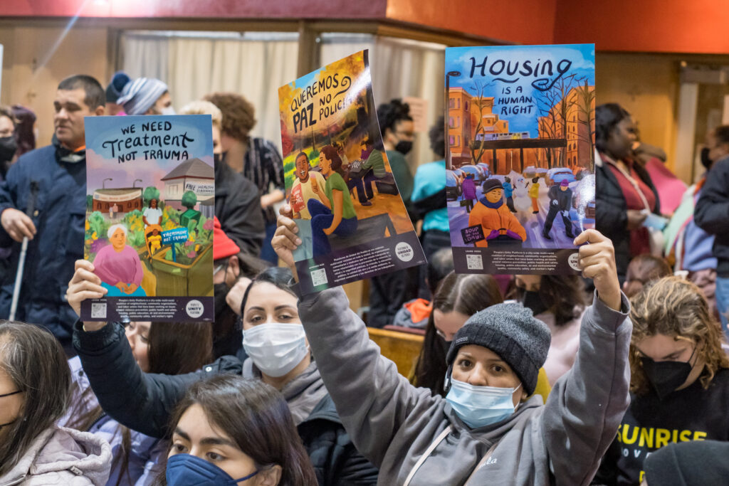 People in a crowded room hold up illustrated signs advocating for treatment over trauma, less police presence, and housing as a human right.