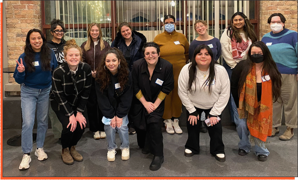A group of fifteen people pose indoors, some standing and others kneeling. Most are smiling; several wear glasses and face masks. Name tags are visible on their shirts.