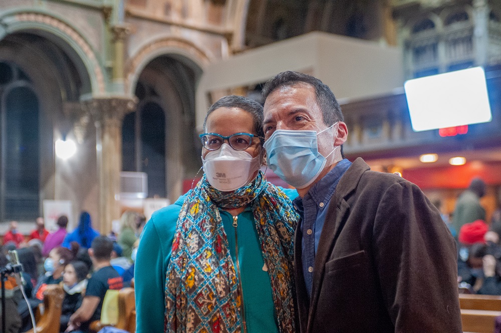 Two people wearing face masks pose for a photo inside a large, ornate building with other people and arches visible in the background.