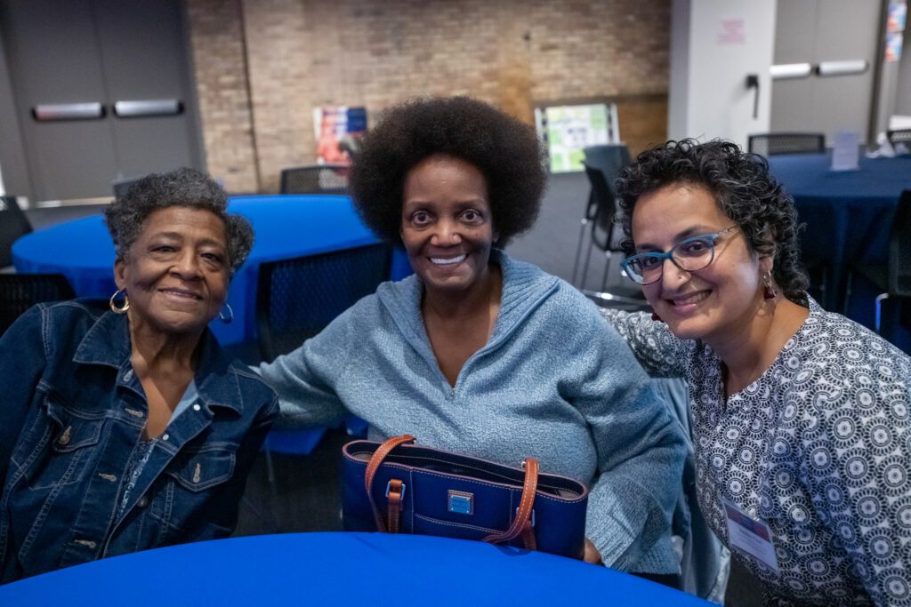 Three people sit together at a blue table, smiling at the camera in an indoor event space with brick and white walls.